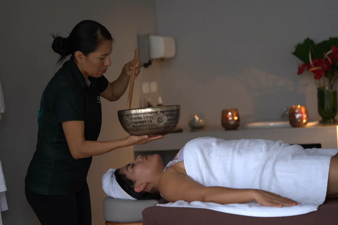 A therapist holding a singing bowl above a guest lying on a treatment table by candlelight