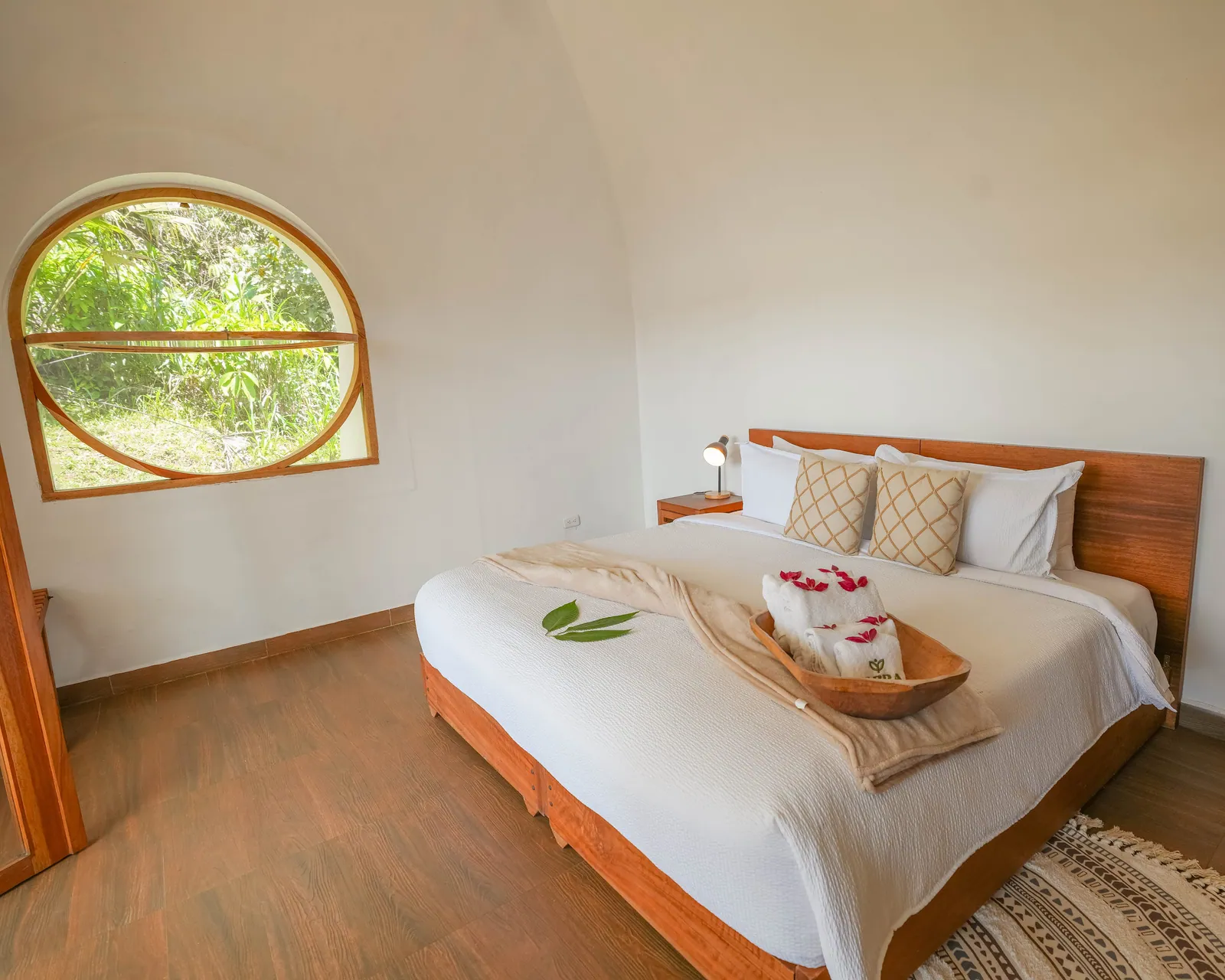 Inside a dome room: a bed with folded towels and leaves laid on the cover, and a round wooden window onto the garden
